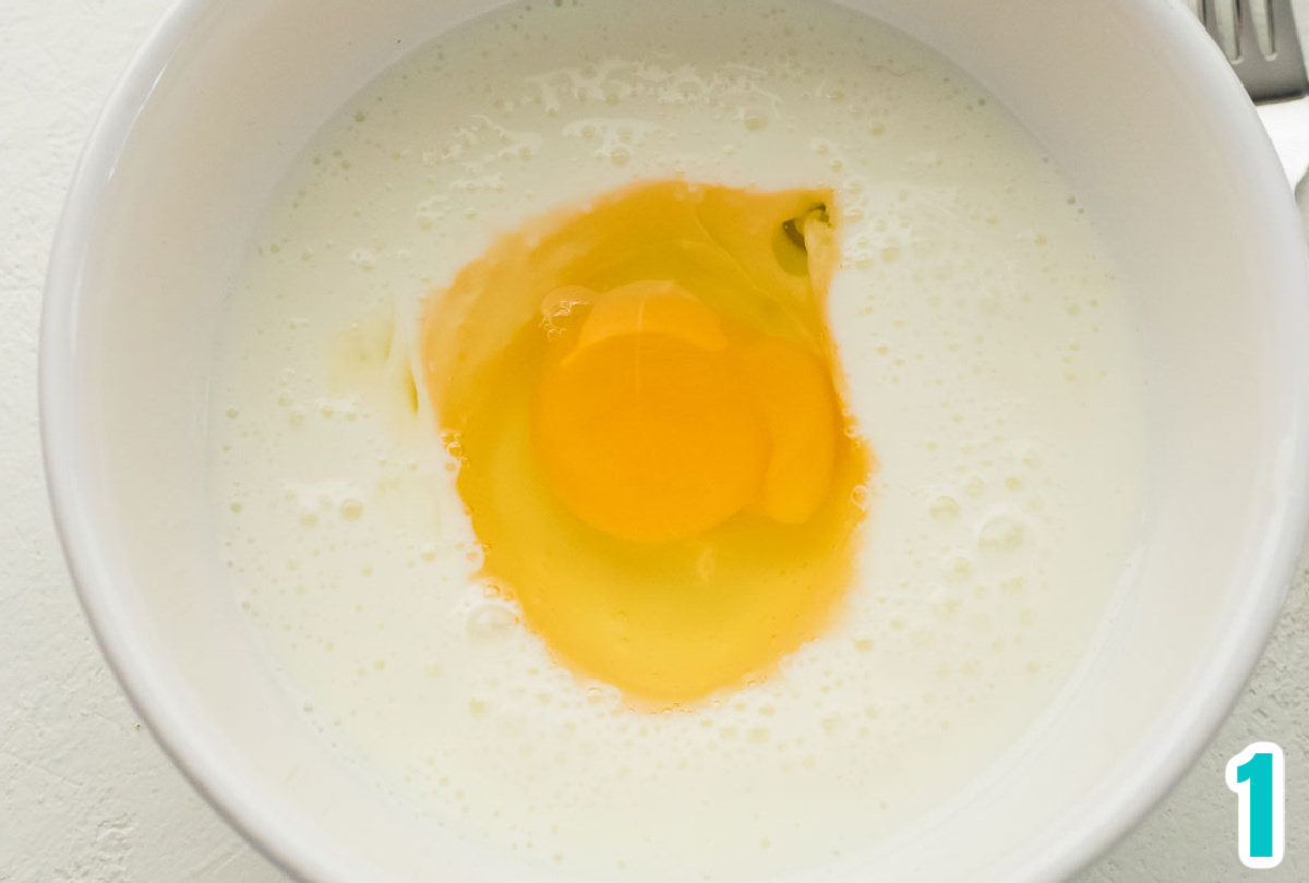 Closeup on a small white bowl filled with buttermilk and a single cracked egg.