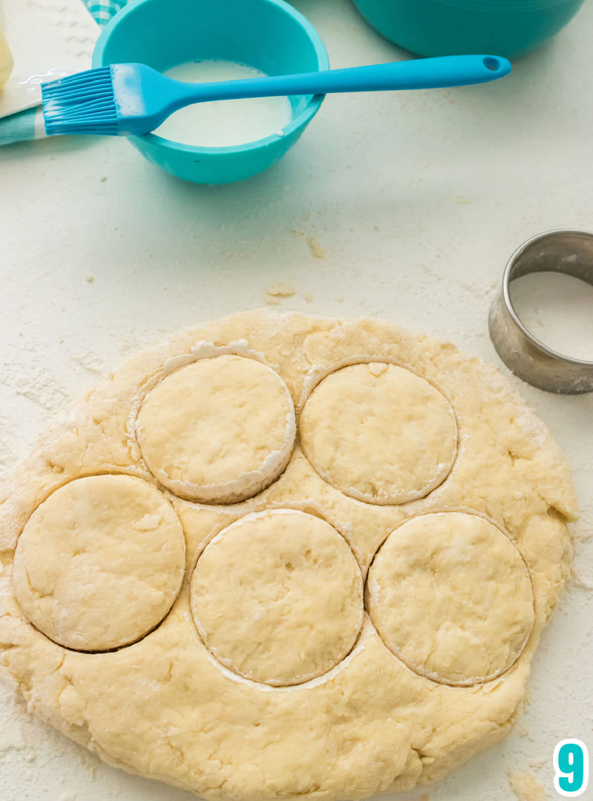Closeup on a circle of Biscuit Dough pressed on a white table with a biscuit cutter and a small bowl filled with milk.
