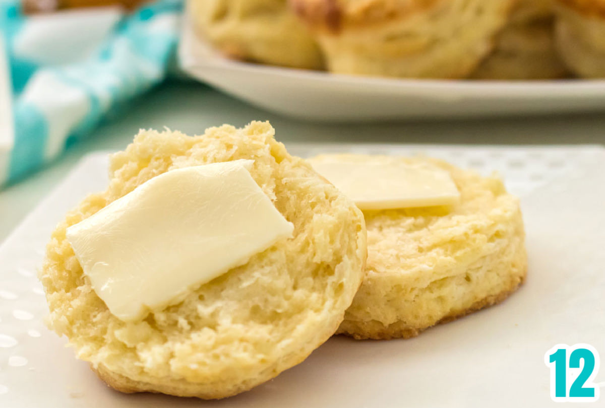 Closeup on a small white plate with a Buttermilk Biscuit that has been cut in half and covered with a pat of butter.