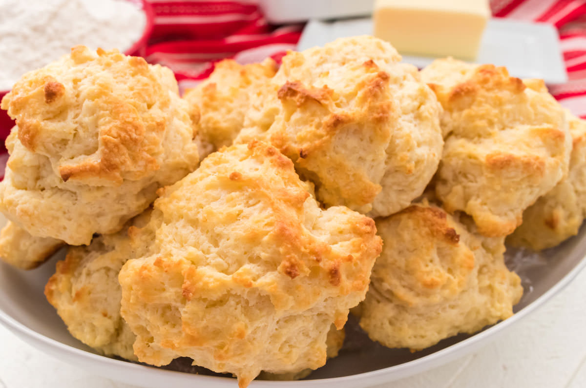 Closeup on a white serving bowl filled with Buttermilk Drop Biscuits sitting in front of a red table linen and a white butter dish.