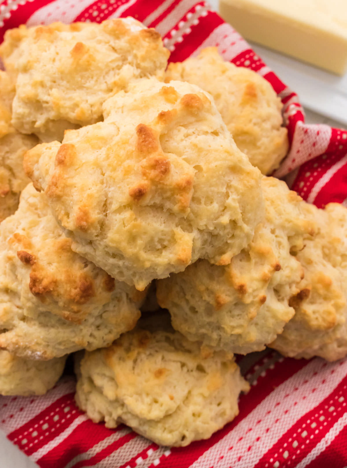 Closeup on a stack of Buttermilk Drop Biscuits in a basket with a red towel next to a butter dish.