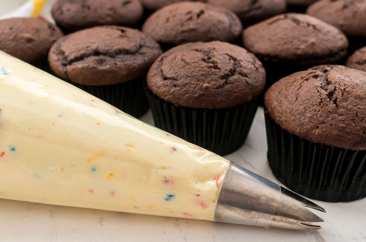 Closeup on a decorating bag filled with Cake Batter Frosting sitting next to a batch of unfrosted chocolate cupcakes.