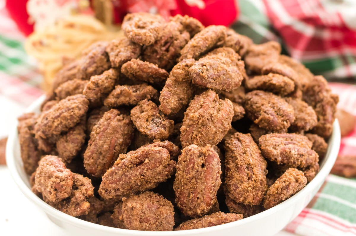 Close up on a white bowl filled with Candied Pecans sitting on a table with Christmas Table linen behind it.