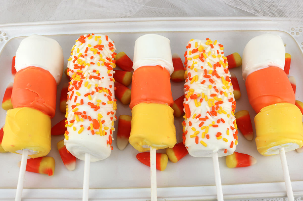 Closeup on five Candy Corn Marshmallow Pops laying on a white serving platter surrounded by real Candy Corn candies.