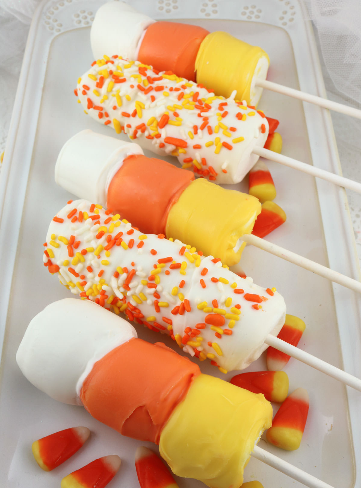Closeup of five Candy Corn Marshmallow Pops laying on a white serving platter on a white table surrounded by Candy Corn candy.