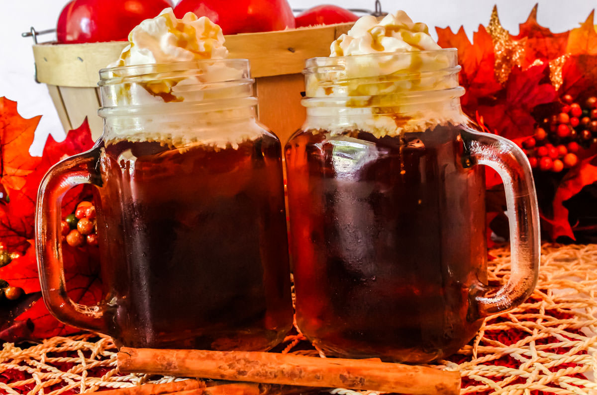 Two glass mugs filled with Caramel Apple Cider topped with Whipped Cream with two cinnamon sticks.