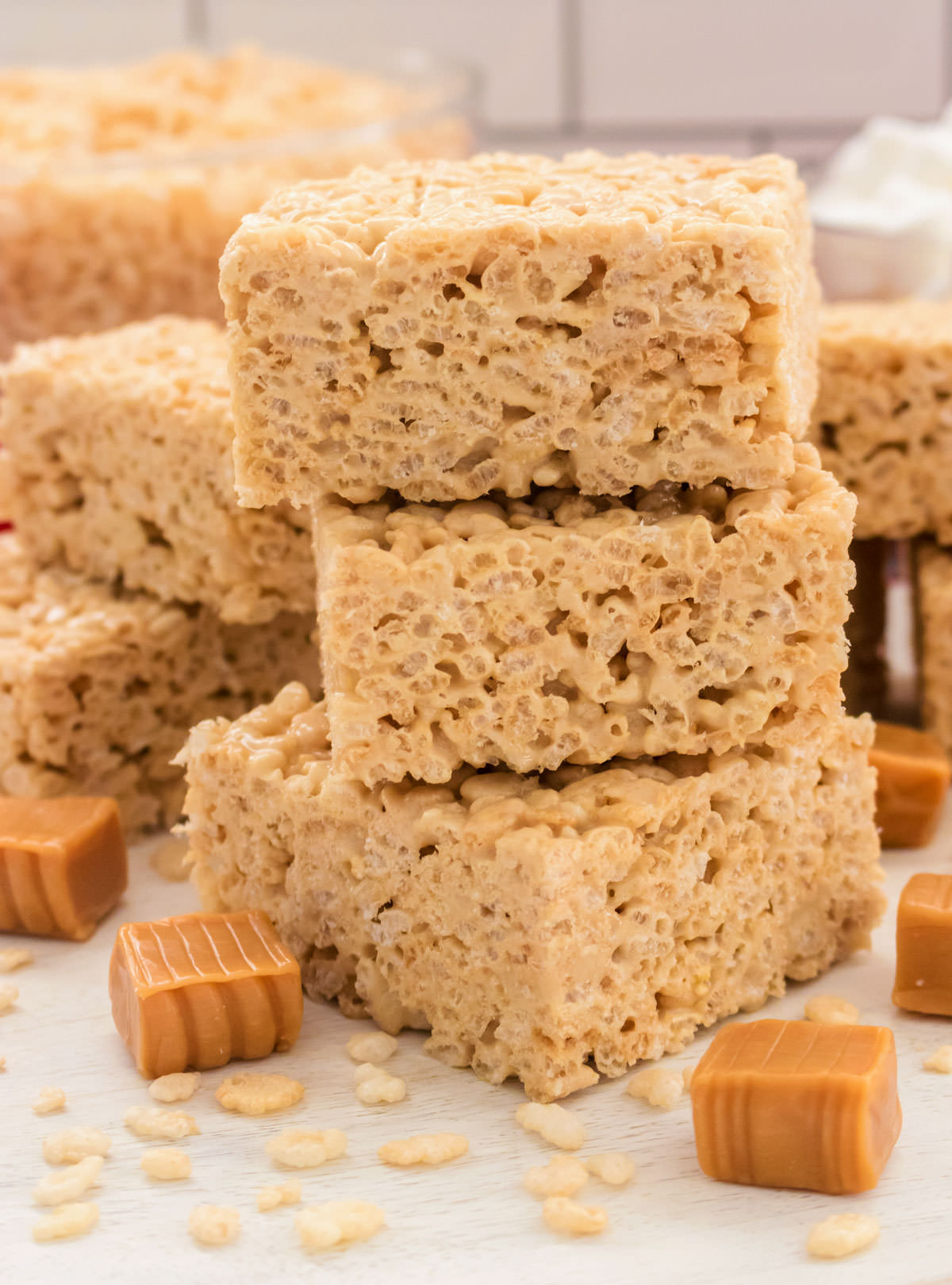 Closeup on a stack of three Caramel Rice Krispie Treats sitting on a white table surrounded by caramel candies and Rice Krispie Cereal.