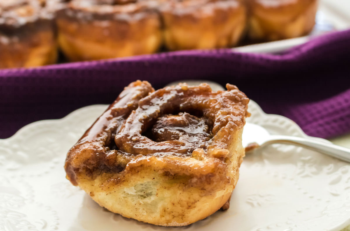 Single Caramel Sticky Bun sitting on a white plate with a purple kitchen towel behind it.