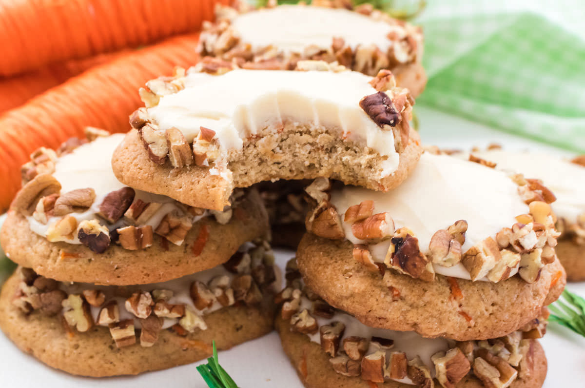 Closeup on a stack of Carrot Cake Cookies with Cream Cheese Frosting sitting on a white surface in front of a green table linen and decorated carrots.