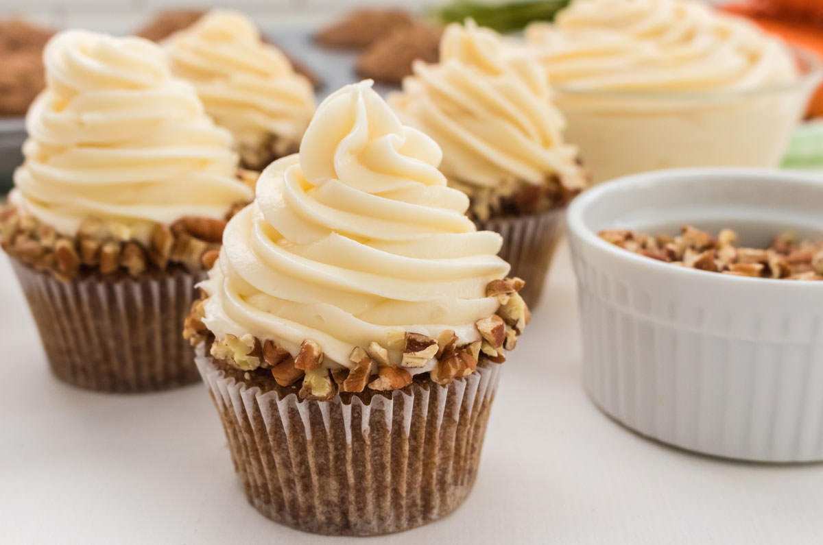 Closeup on four Carrot Cake Cupcakes frosted with Cream Cheese Frosting sitting on a white table in front of a bowl of cream cheese frosting, a cupcake tin full of cupcakes and a ramekin full of nuts.