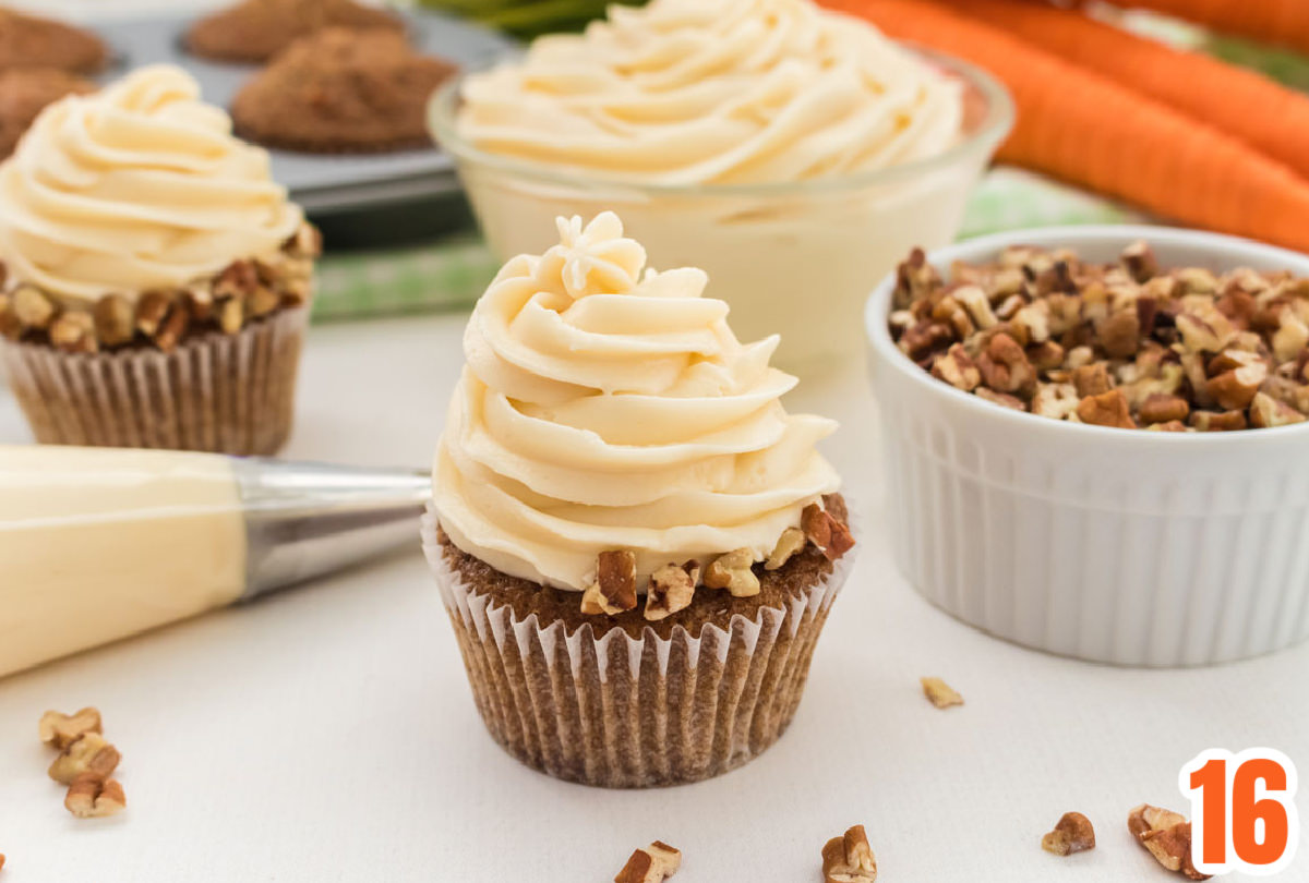 Carrot Cake Cupcake sitting on a white table next to a decorating bag filled with cream cheese frosting, a glass bowl filled with frosting and a ramekin filled with chopped nuts.