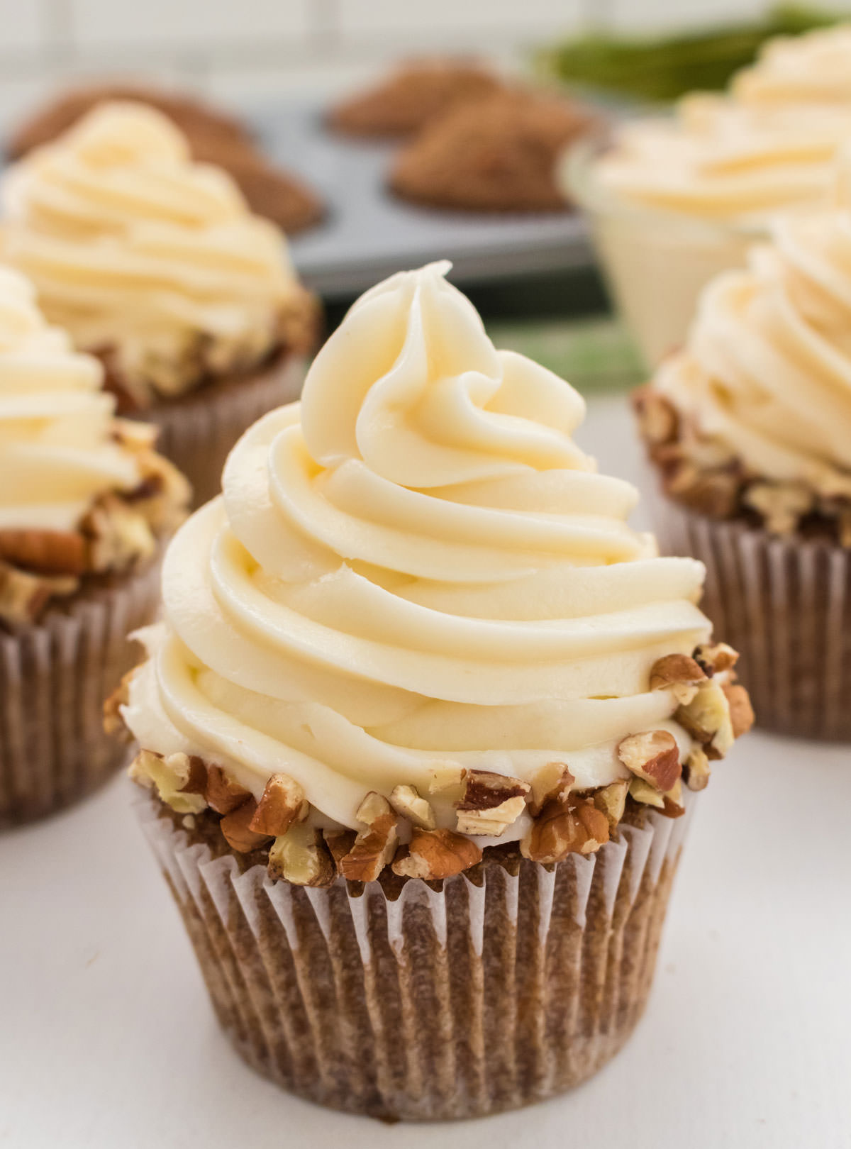 Closeup on a collection of homemade Carrot Cake Cupcakes sitting on a white table in front of a cupcake tin filled with unfrosted carrot cake cupcakes.