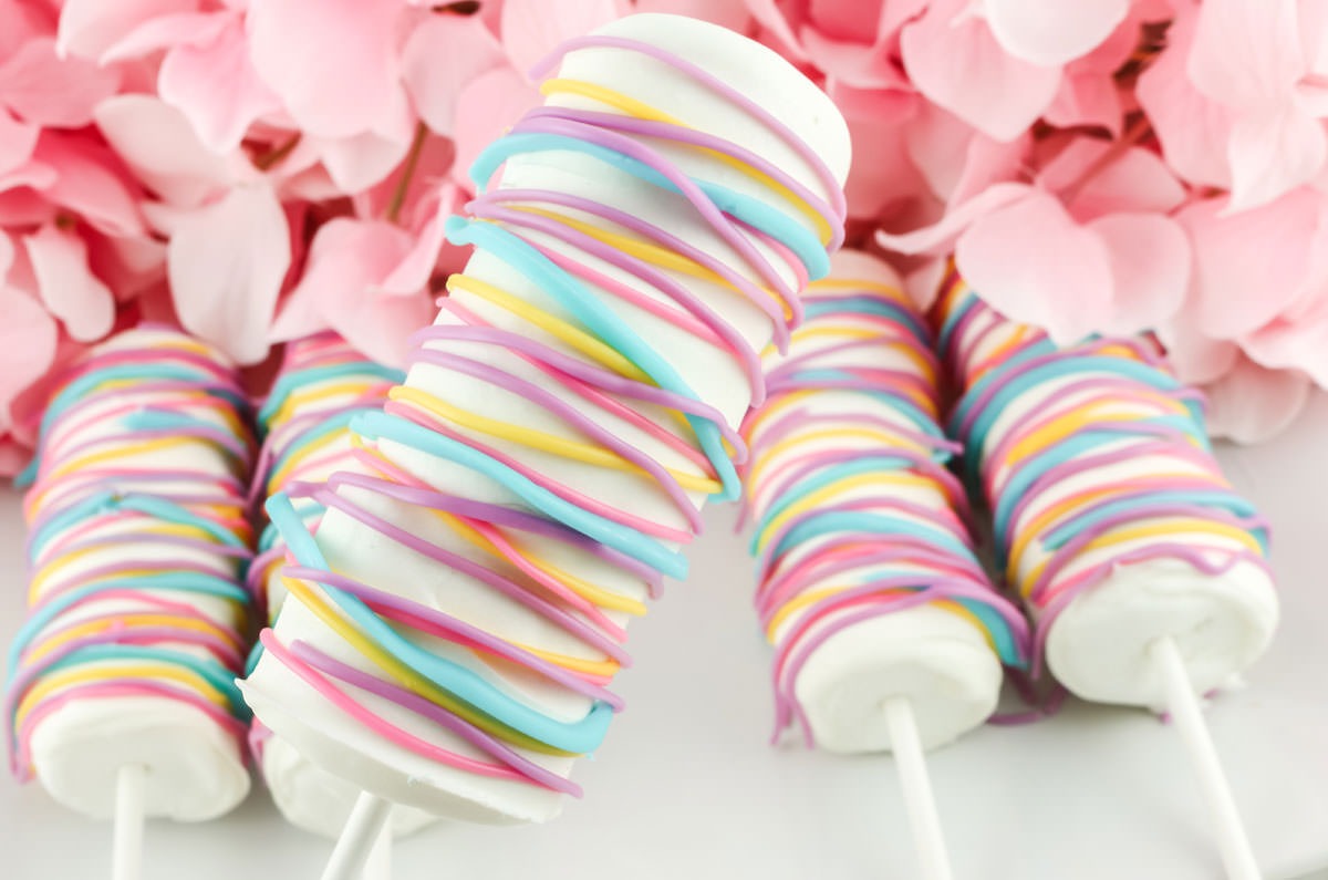 Close up of a Celebration Marshmallow Pop being held up over a white platter covered with more marshmallow pops.