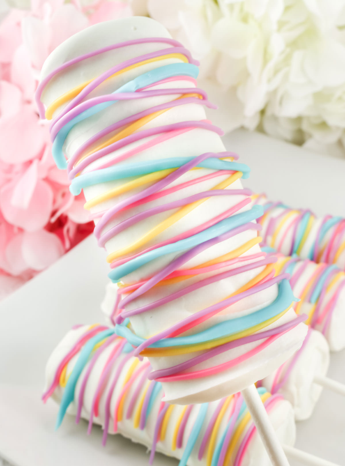 Close up of a White Chocolate Celebration Marshmallow Pop being held up by the lollipop stick over a plate of other pops.