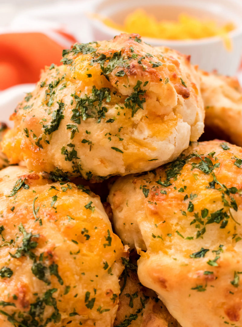 A plateful of Cheesy Garlic Biscuits sitting on a table in front of a ramekin full of shredded cheddar cheese.