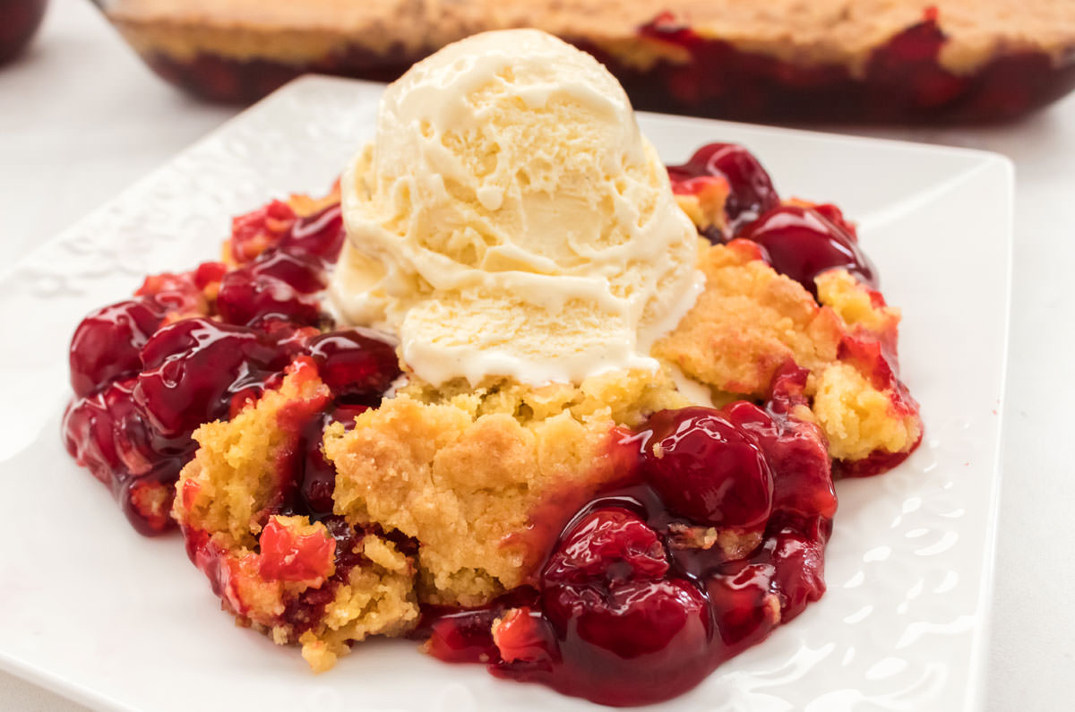 Closeup on a piece of Cherry Dump Cake topped with a scoop of vanilla ice cream sitting in front of the pan of Dump Cake.