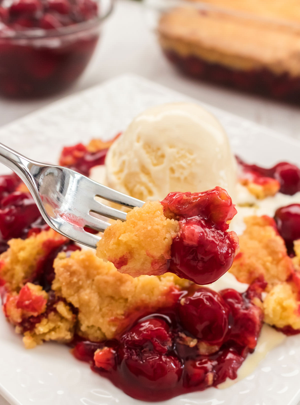 Closeup on a white plate filled with Cherry Dump Cake topped with a scoop of vanilla ice cream and a fork holding a piece of dump cake.