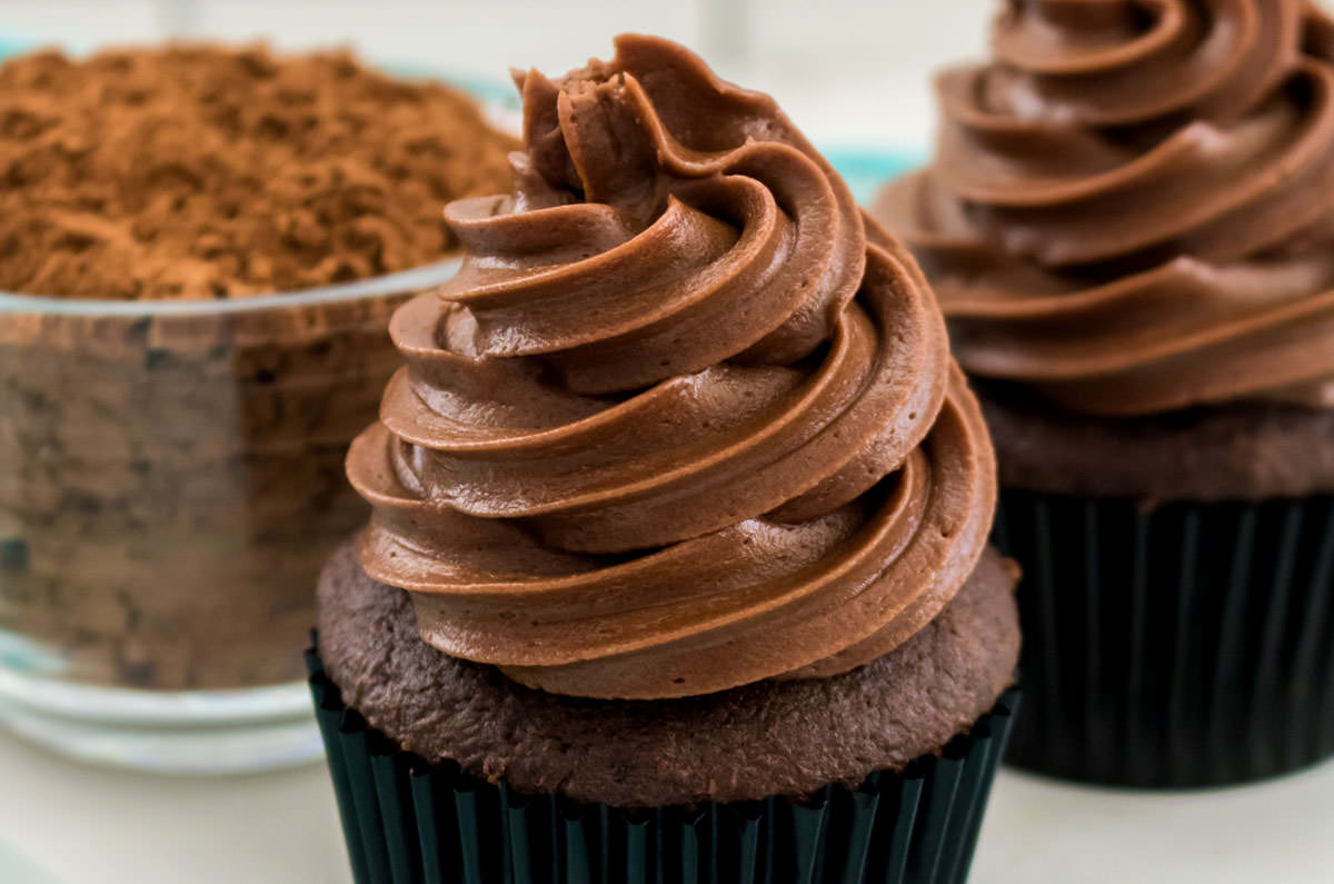 Closeup on two cupcakes sitting on a white table frosted with Chocolate Cream Cheese Frosting.