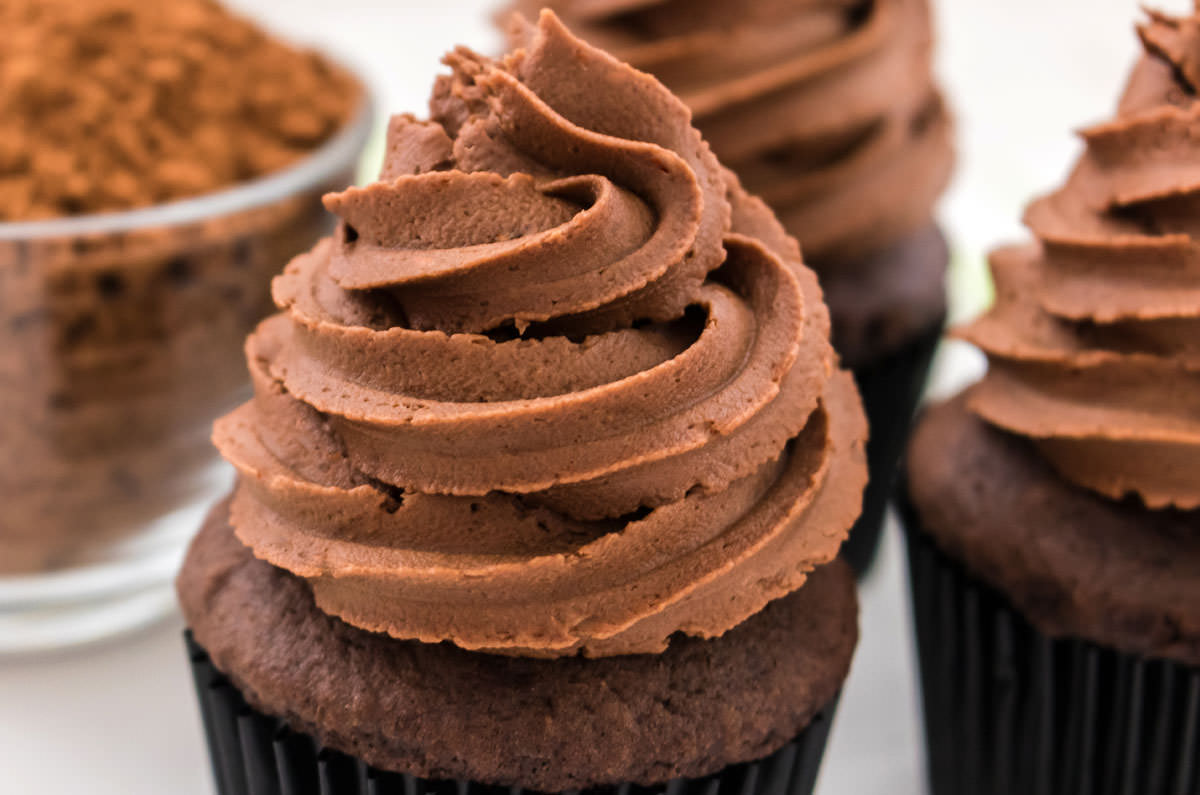 Closeup on three cupcakes sitting on a white table topped with The Best Chocolate Mint Buttercream Frosting next to a glass bowl filled with cocoa powder.