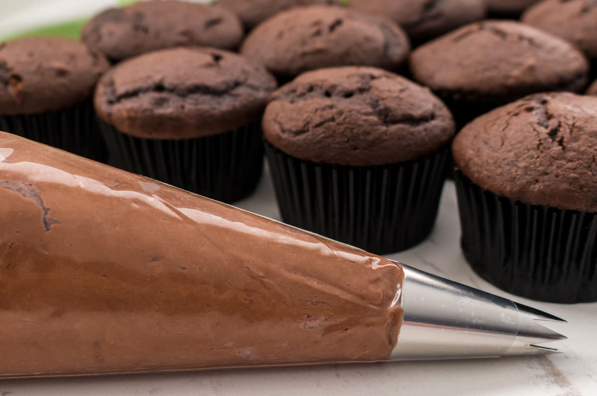 Closeup on a decorating bag filled with The Best Chocolate Mint Buttercream Frosting sitting on a white table in front of a batch of unfrosted cupcakes.