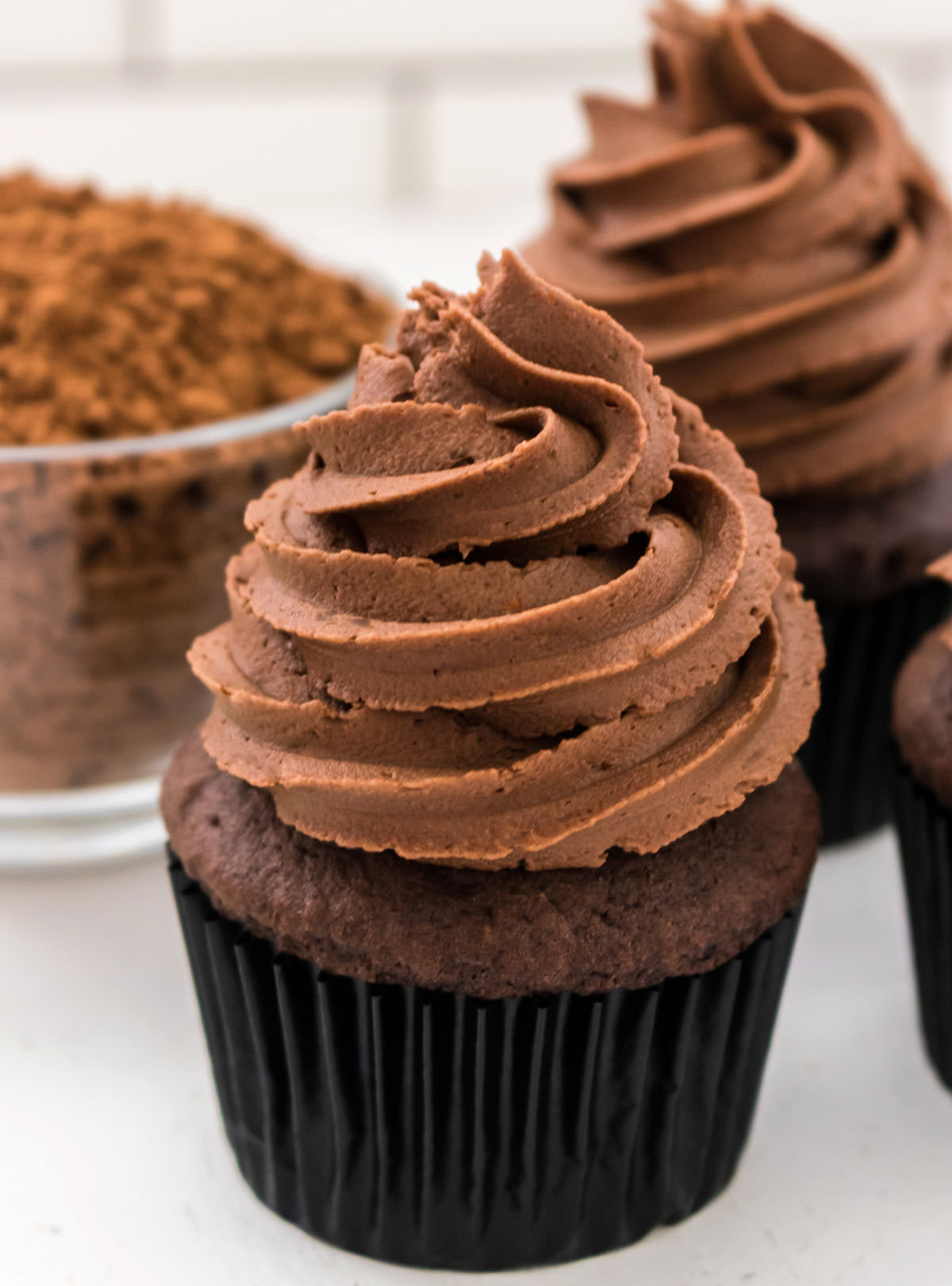 Closeup on a chocolate cupcake topped with The Best Chocolate Mint Buttercream Frosting sitting next to a glass bowl filled with cocoa powder.