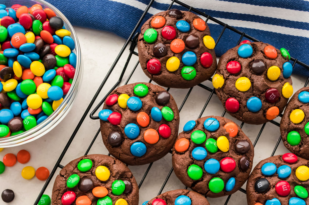 Overhead shot of a batch of Chocolate M&M Cookies sitting on a cooling rake sitting next to a bowl of M&M Candies.