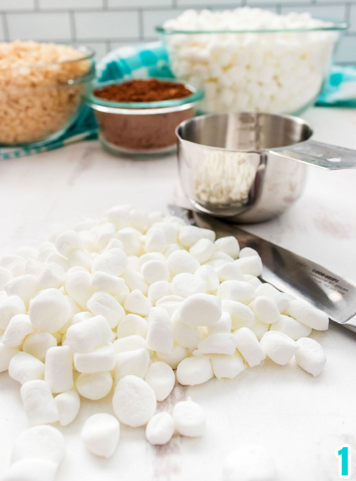 Chopped up Mini Marshmallows laying on a white table sitting next to a measuring cup, and bowls of cocoa powder, marshmallows and Rice Krispie Cereal.