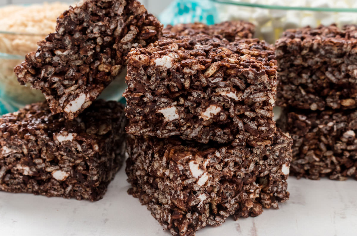 Closeup on a stack of six Chocolate Rice Krispie Treats sitting on a white table in front of bowls of cereal and marshmallows.