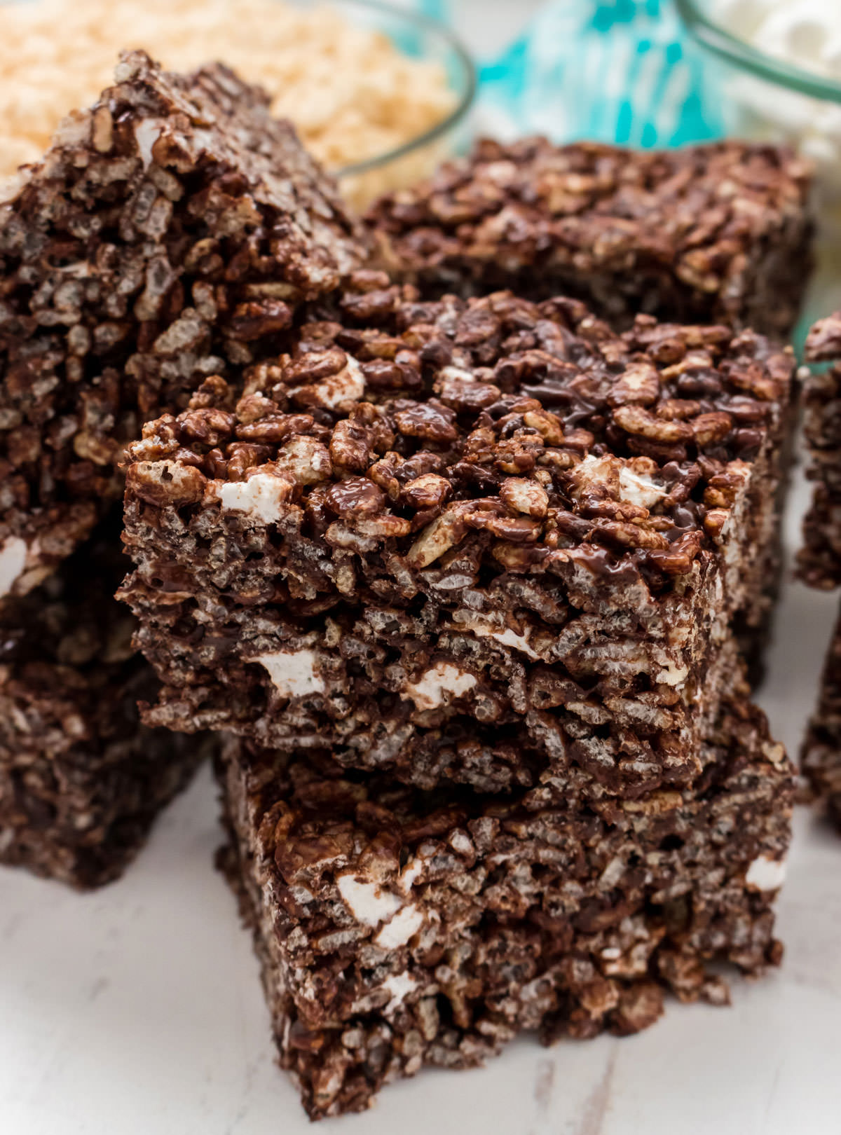 Closeup on a stack of Chocolate Rice Krispie Treats sitting on a white surface in front of a glass bowl filled with Rice Krispie Cereal.