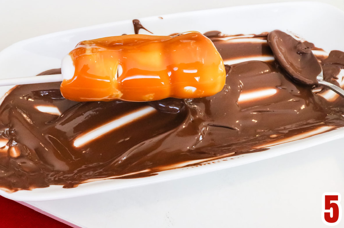 Closeup on a caramel covered marshmallow pop being covered by melted dark chocolate.