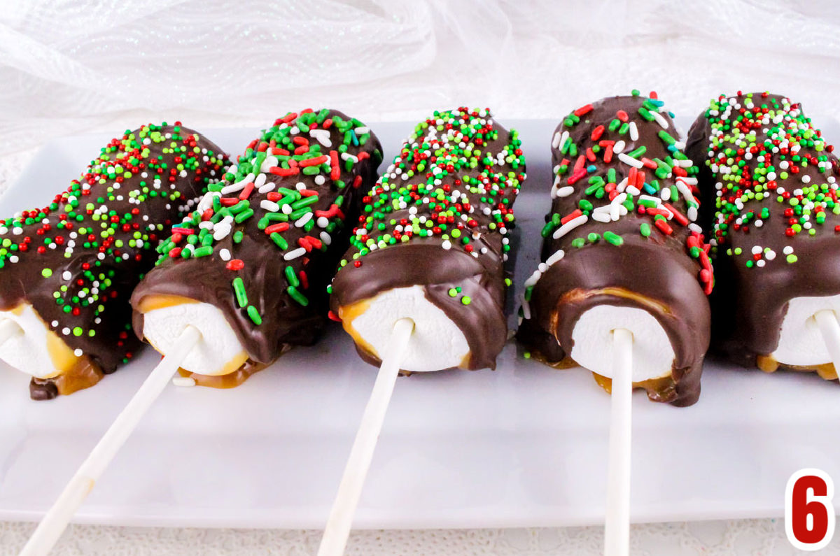 Sideview of five Christmas Caramel Marshmallow Pops laying on a white plate on a white table.