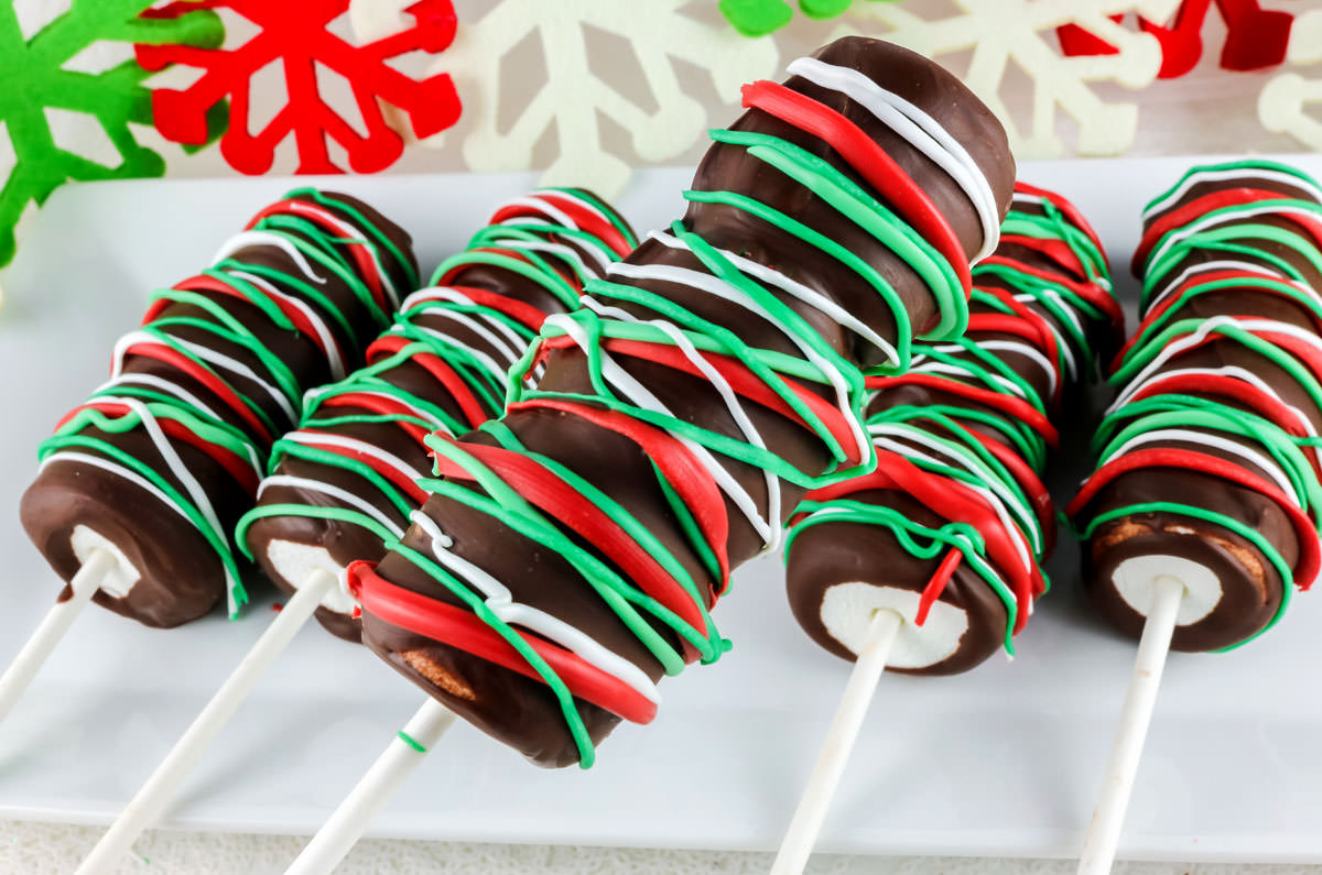 Closeup on five Christmas Marshmallow Pops laying on a white serving platter in front of Christmas Decorations.