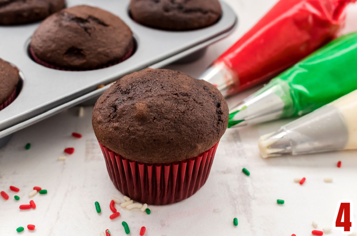 Chocolate cupcake in a red cupcake liner sitting next to three decorating bags filled with red, green and white frosting.