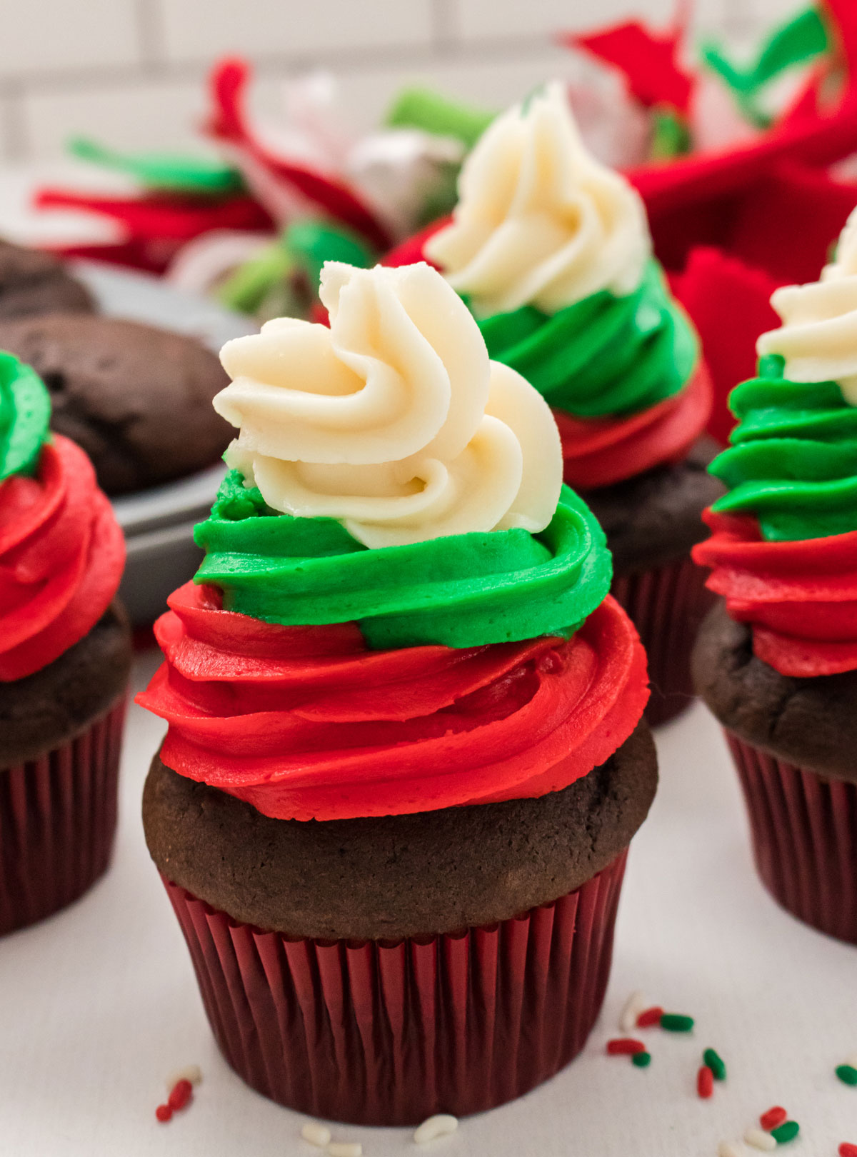 Closeup on four Easy Christmas Cupcakes sitting on a white table in front of a tin of chocolate cupcakes and some Christmas decorations.
