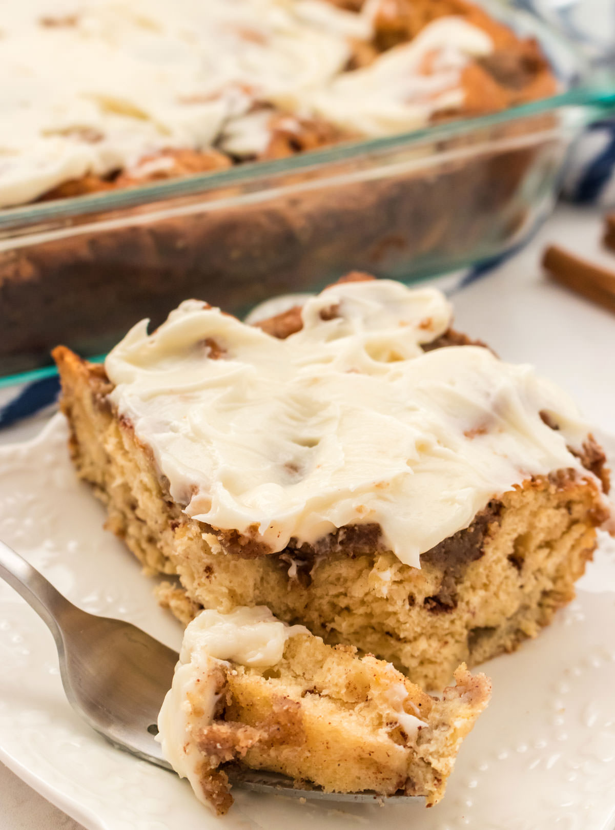 Close up of a piece of Cinnamon Roll Breakfast Casserole sitting on a white plate in front of the pan of remaining casserole.