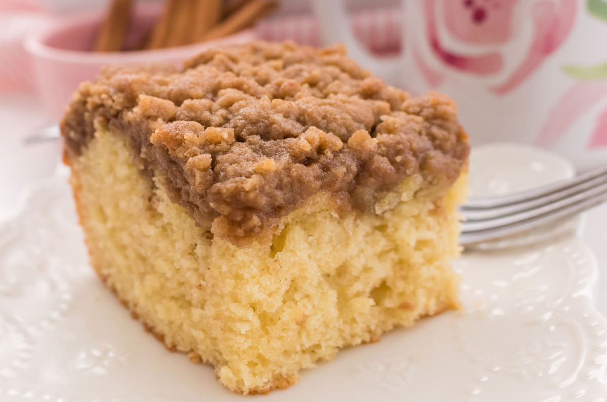 Close up of a piece of Coffee Cake on a white plate along with a flowered coffee mug and a ramekin filled with Cinnamon sticks.