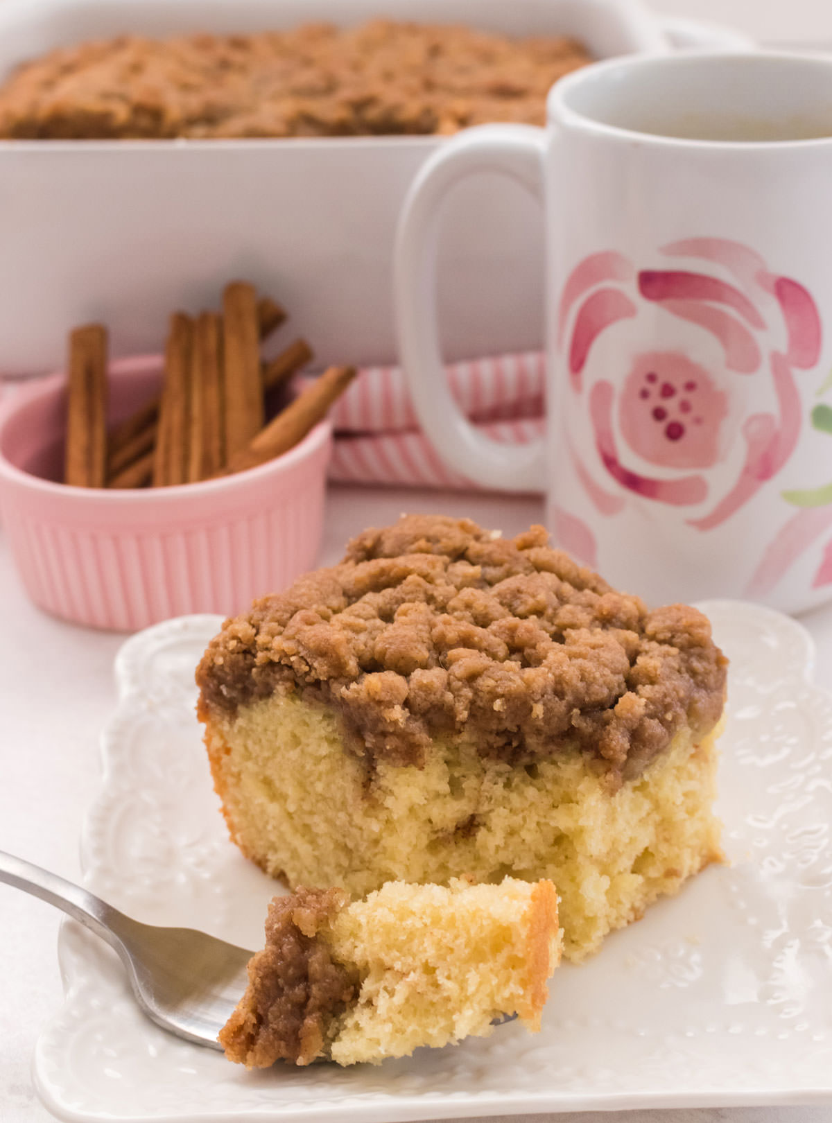 Close up of a piece of Classic Coffee Cake sitting on a white plate in front of a cup of coffee and the rest of the cake.