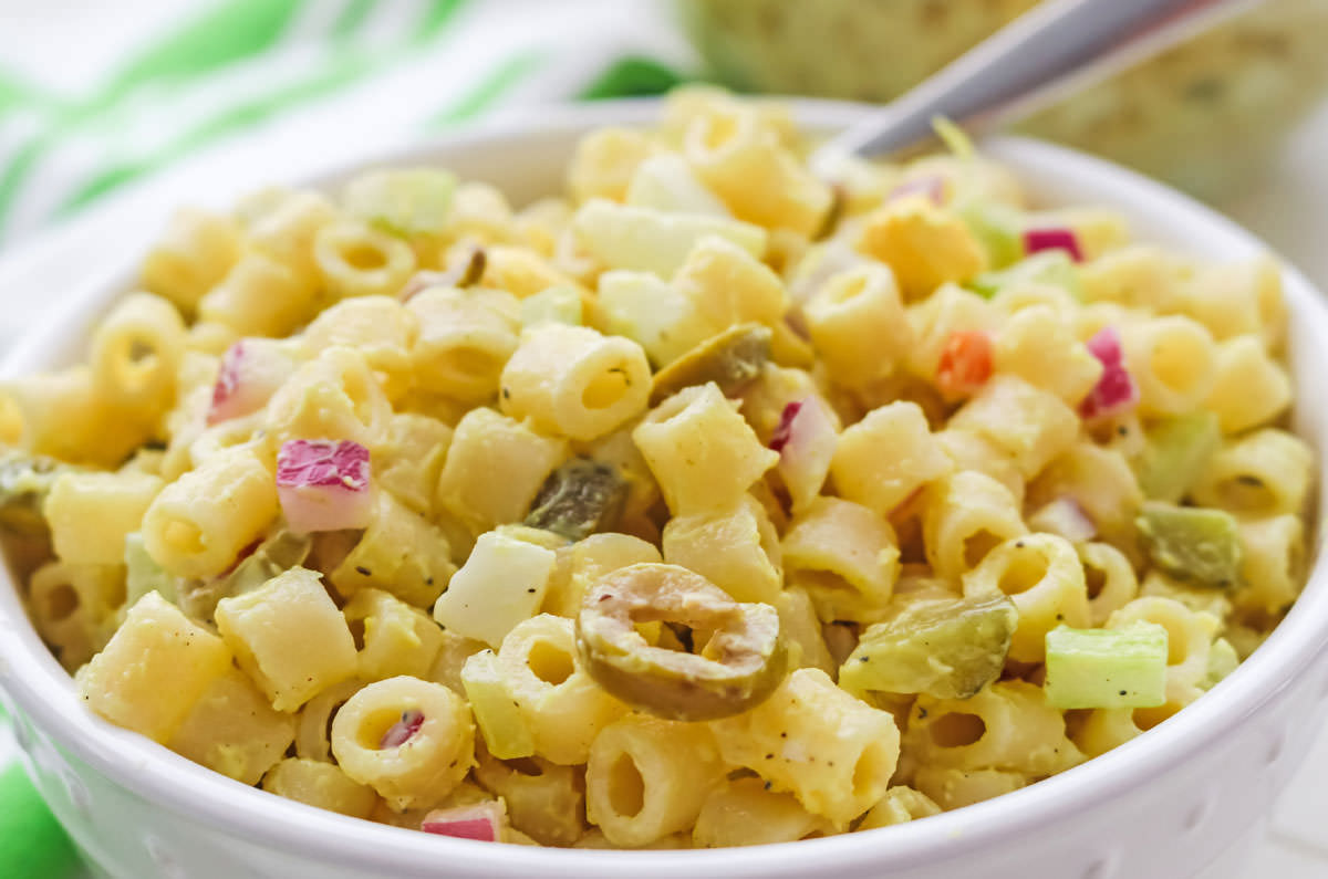 Closeup on a white bowl filled with Macaroni Salad and a silver spoon in front of a green and white towel.