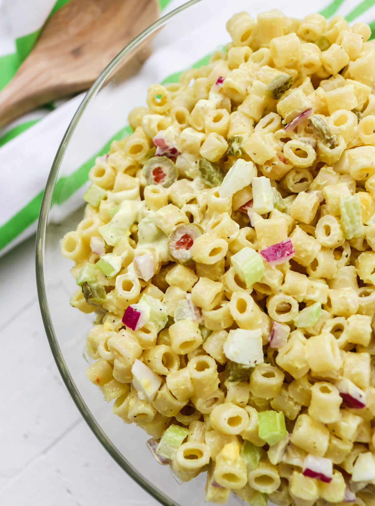 Closeup on a clear glass serving bowl filled with Classic Macaroni Salad sitting on a white table with a towel and a wooden spoon in the background.