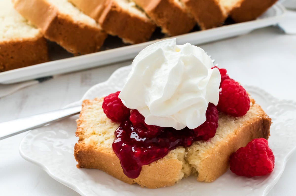 A piece of pound cake, covered in raspberries and whipping cream, sitting on a white plate in front of the remaining pieces of cake.