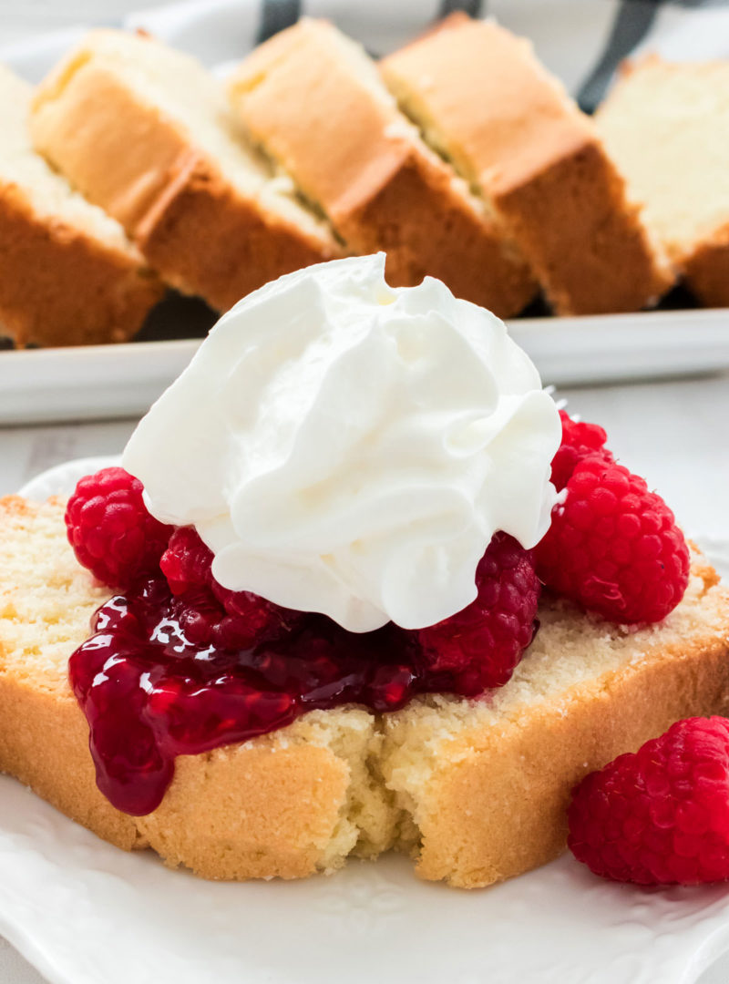 Close up of a piece of Classic Pound Cake topped with fresh Raspberries and whipped cream, more pound cake featured in the background.