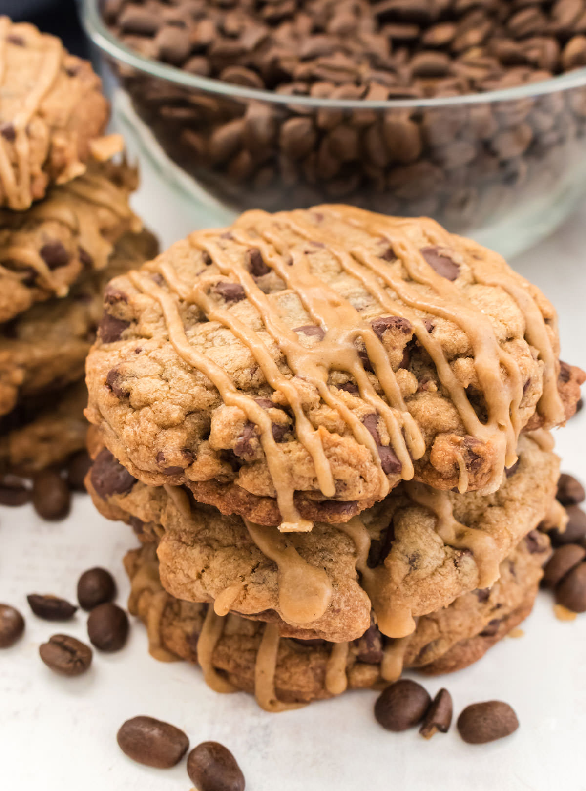 Closeup on a stack of three Coffee Chocolate Chip Cookies sitting on a white table surrounded by more cookies and a glass bowl filled with coffee beans.