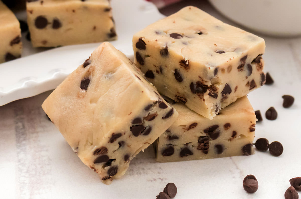 Closeup on three pieces of Cookie Dough Fudge sitting on a white surface surrounded by Mini Chocolate Chips.