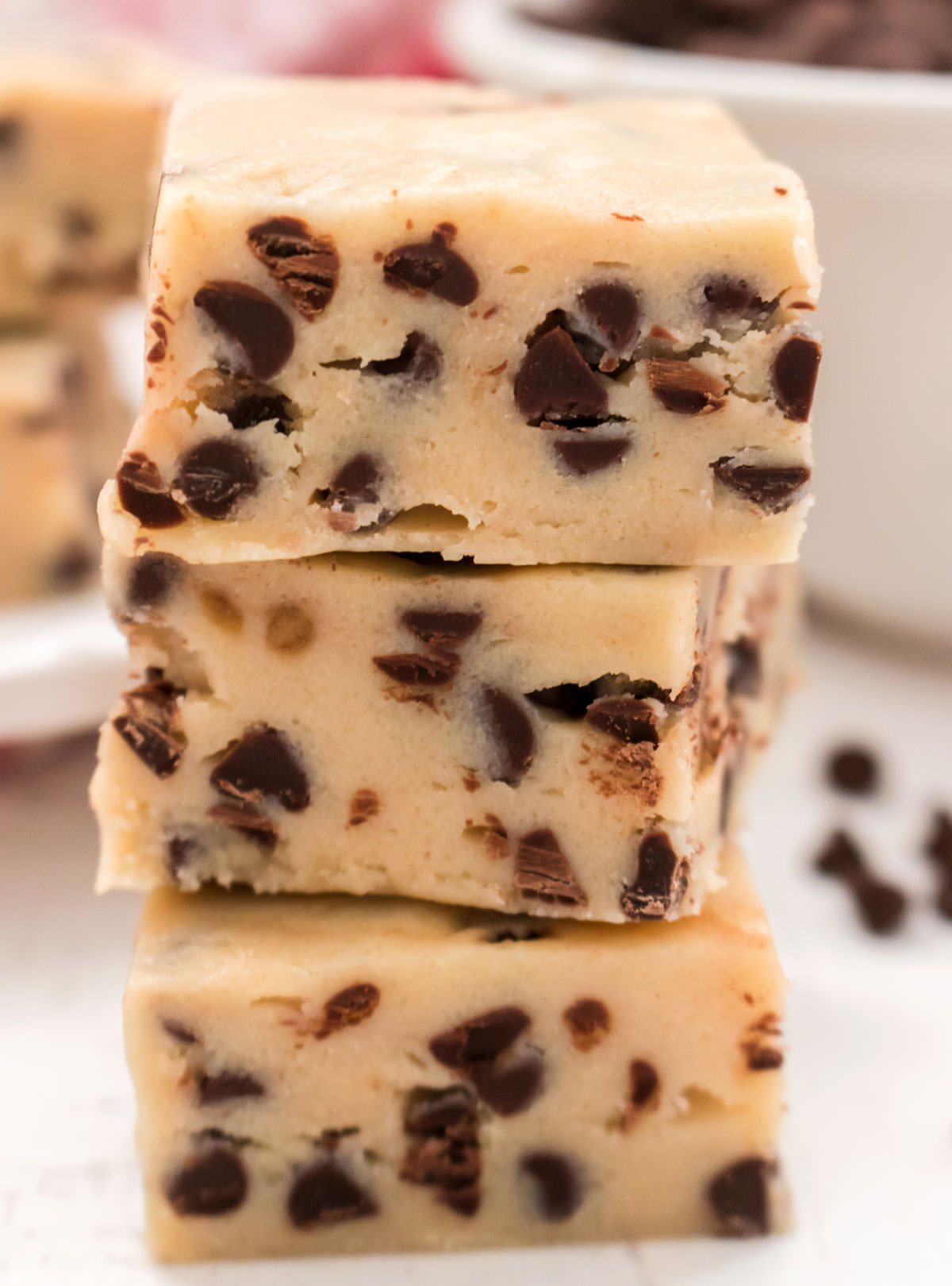 Closeup on a stack of three Cookie Dough Fudge candies sitting on a white surface in front of a plate full of more fudge.
