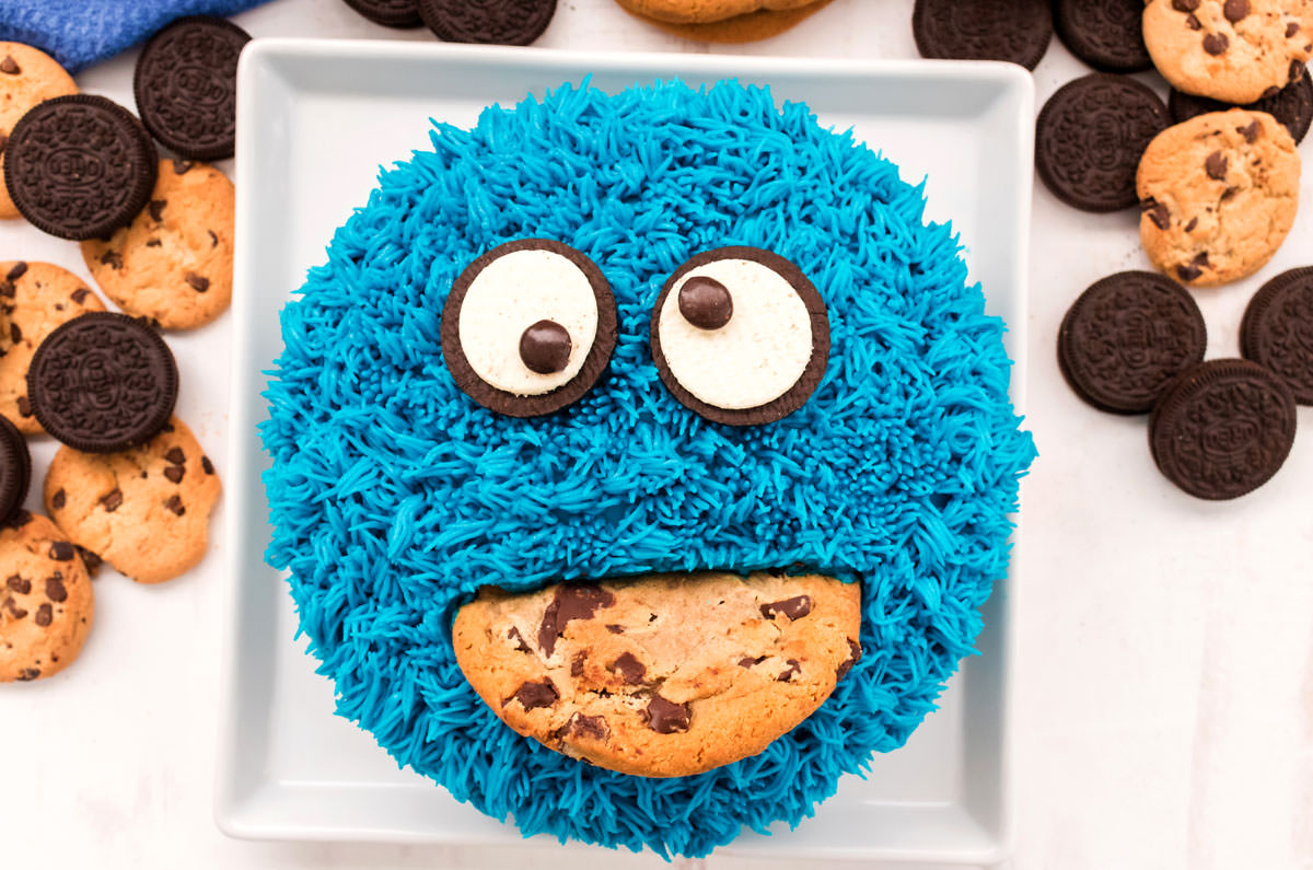 Closeup of a Cookie Monster Cake sitting on a white square plate which is sitting on a white surface surrounded by chocolate chip cookies and Oreo Cookies.
