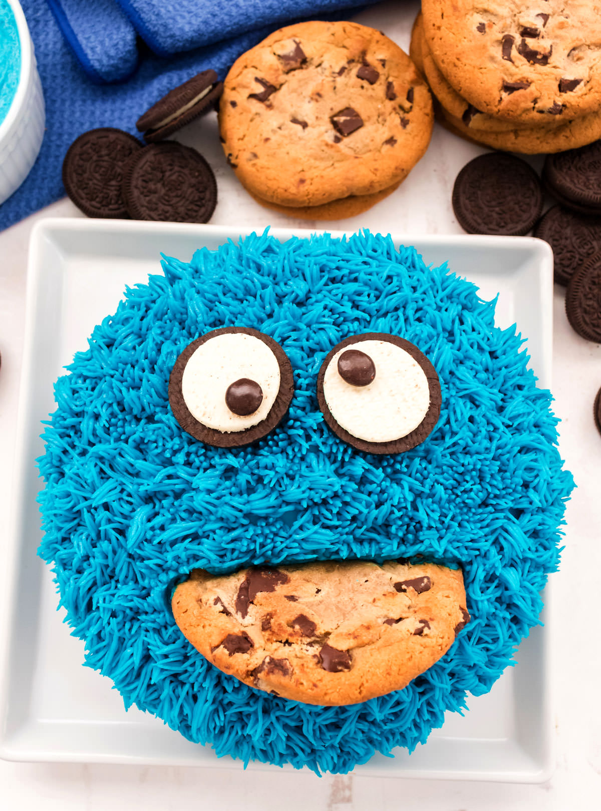 Closeup on a Cookie Monster Cake sitting on a white cake plate on a white table surrounded by Oreo and Chocolate Chip Cookies.