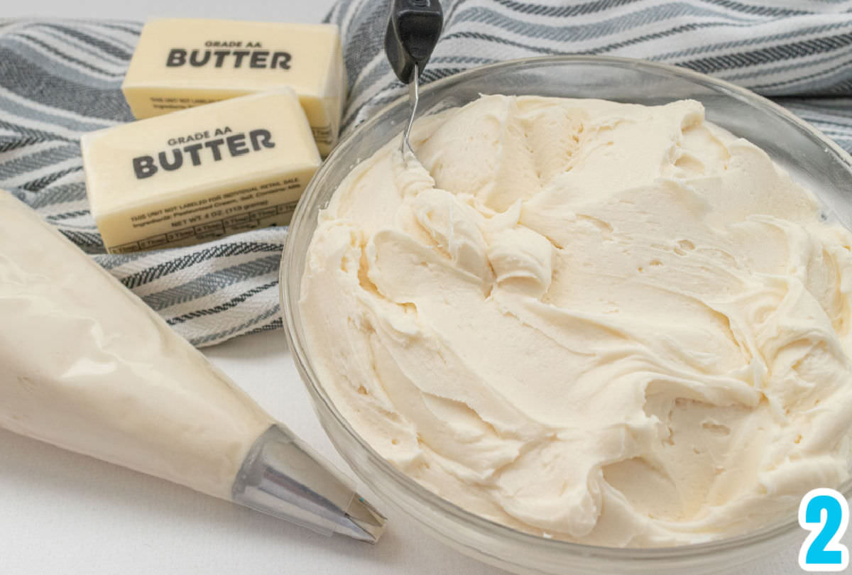 Clear glass bowl filled with homemade buttercream frosting sitting next to two sticks of butter and a decorating bag filled with frosting.