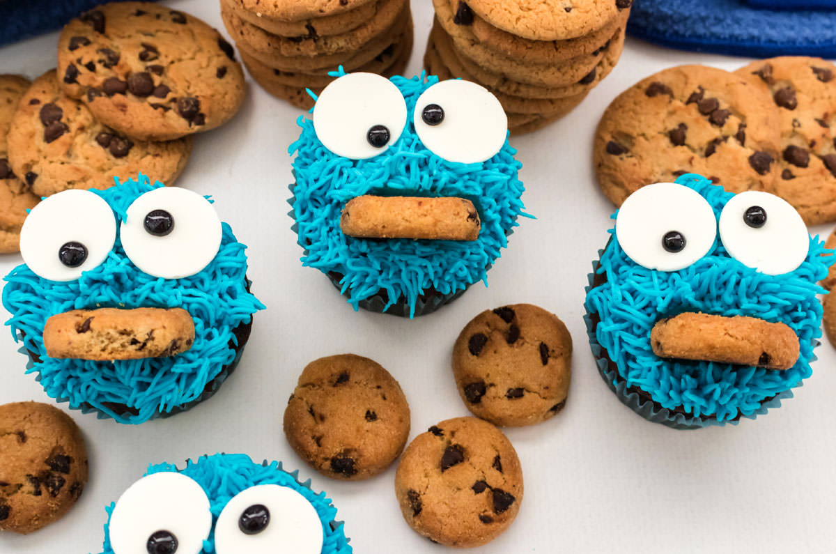 Three Cookie Monster Cupcakes sitting on a white surface surrounded by chocolate chip cookies.