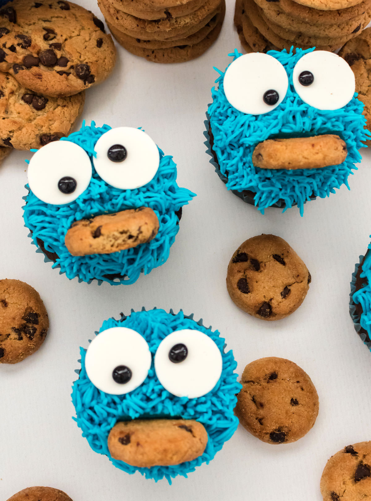 Three Cookie Monster Cupcakes sitting on a white table surrounded by Chocolate Cookies.