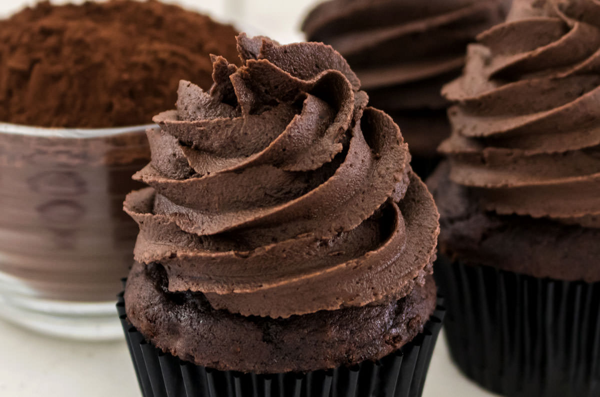 Closeup on three chocolate cupcakes topped with The Best Dark Chocolate Buttercream Frosting sitting next to a glass bowl filled with cocoa powder.